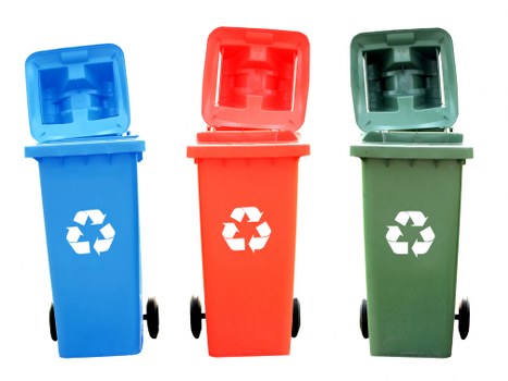 The image displays three wheeled waste bins positioned side by side on a paved surface. The bin on the left is blue with a partially open lid, revealing a smooth plastic interior. It features a white recycling symbol on its front face and has small black wheels at the base. The middle bin is red, also with a partially open lid showing a similar interior, and has a white recycling symbol on the front. The right bin is green, with its lid slightly tilted backward, exposing the interior, and similarly bears a white recycling symbol. All three bins are standard-sized for domestic or commercial waste collection, placed outdoors in what appears to be a driveway or pavement area, with a plain background and natural lighting. This setup is consistent with rubbish and recycling collection services such as those offered by Commercial Waste Removal East Ham, serving areas within East London postcodes, including nearby locations. The image emphasizes proper waste segregation and disposal practices using clearly color-coded bins.