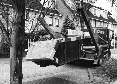 A black tow truck equipped with a hydraulic lift is parked on a residential street, lifting a large open-top metal skip filled with mixed rubbish, including various debris and construction materials. The skip, made of metal with a dull, weathered finish, is suspended in mid-air, partly leaning toward the curb as it is being removed from a driveway. The street is lined with multi-story houses featuring brick and siding exteriors, with trees and shrubs visible along the pavement, indicating a typical suburban area. The scene appears to be in daylight with overcast conditions, casting soft shadows. This image relates to rubbish removal services provided by Commercial Waste Removal East Ham, serving the East Ham area with reliable rubbish disposal for domestic or commercial waste in the local postcode vicinity.