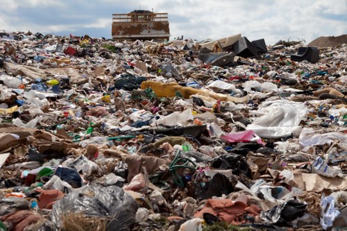 A vast expanse of mixed household and industrial waste spread across an open area, featuring plastic bags, cardboard, paper, broken furniture, and discarded packaging material. The rubbish is scattered unevenly on the ground, creating a dense and cluttered terrain. In the background, a partially constructed or abandoned building can be seen on the horizon, with a partly cloudy sky overhead. The scene suggests an environment awaiting waste collection or clearance, typical of rubbish removal services provided in East Ham. The materials vary in texture, with some plastic items appearing crumpled or torn, while cardboard is flattened or bent. The colour palette includes shades of white, brown, black, and various bright hues from plastic packaging, contributing to the disorderly appearance of the site. This image exemplifies the type of waste that a commercial waste removal service like Commercial Waste Removal East Ham might handle, illustrating the need for professional rubbish clearance to restore cleanliness and environmental standards in the local area.