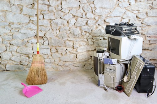 The image shows a cluttered corner of a room or storage area with a stone wall as the background. In the foreground, there is a pink plastic dustpan and a beige broom with a yellow handle leaning against the wall. On the right side, a collection of electronic waste and old computer equipment is piled together, including a beige CRT monitor, an older computer tower, a tower speaker, a keyboard, a mouse, a small white device, and various cables and power cords draped over and around the items. The items appear to be discarded or awaiting rubbish removal, and the setting is a typical space where waste and unwanted items are temporarily stored before disposal or collection. This scene represents typical household or small business electronic waste ready for rubbish removal services, such as those offered by Commercial Waste Removal East Ham, in the East London area postcode region.